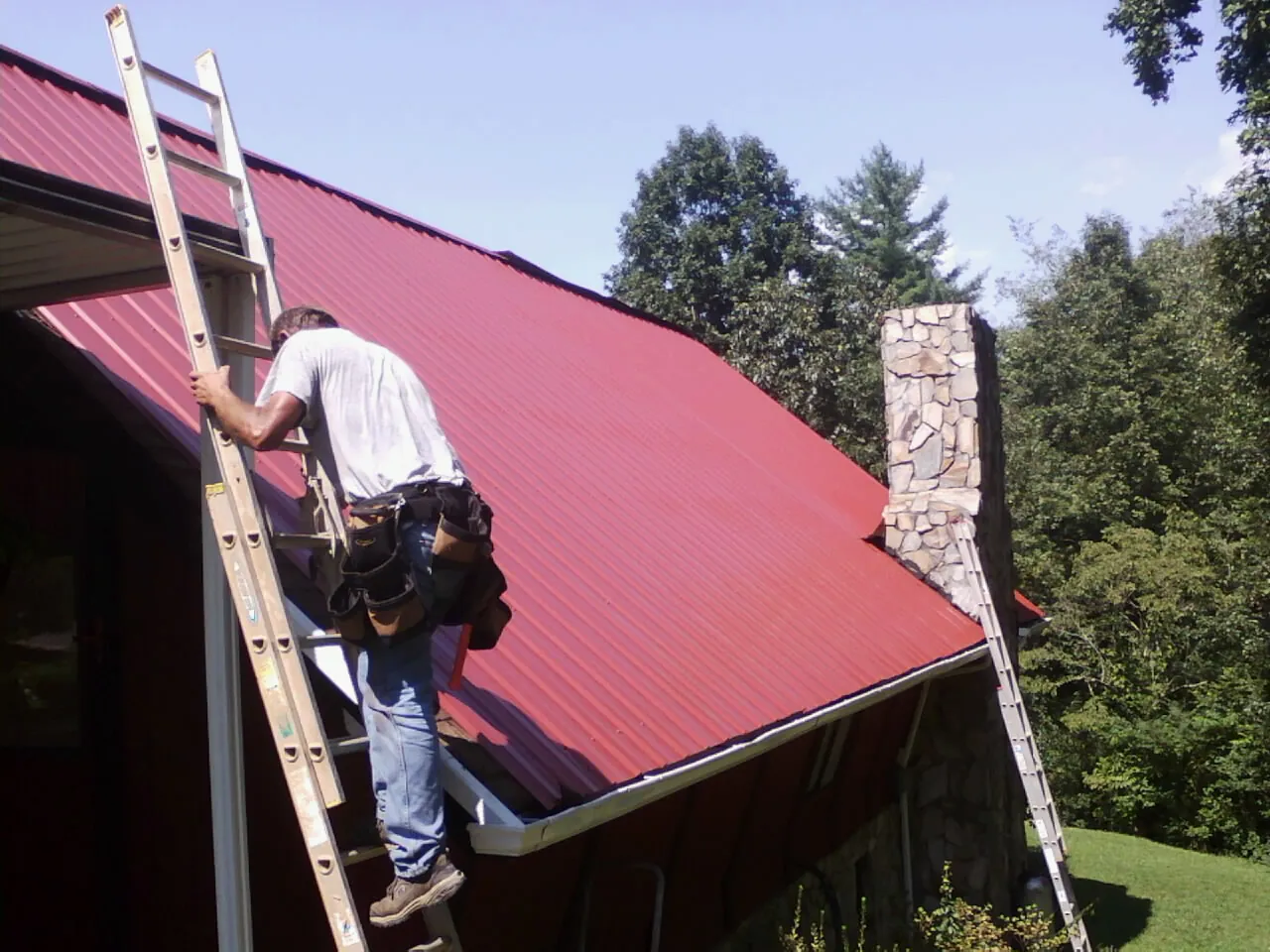 Roofer on a ladder beside a red metal roof and stone chimney — Soffit & Fascia Repair by Roofing Brothers in Wattsburg